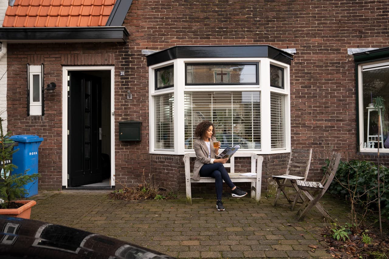 Voorkant huis, niet helemaal in beeld, vrouw op bankje met brochure.
