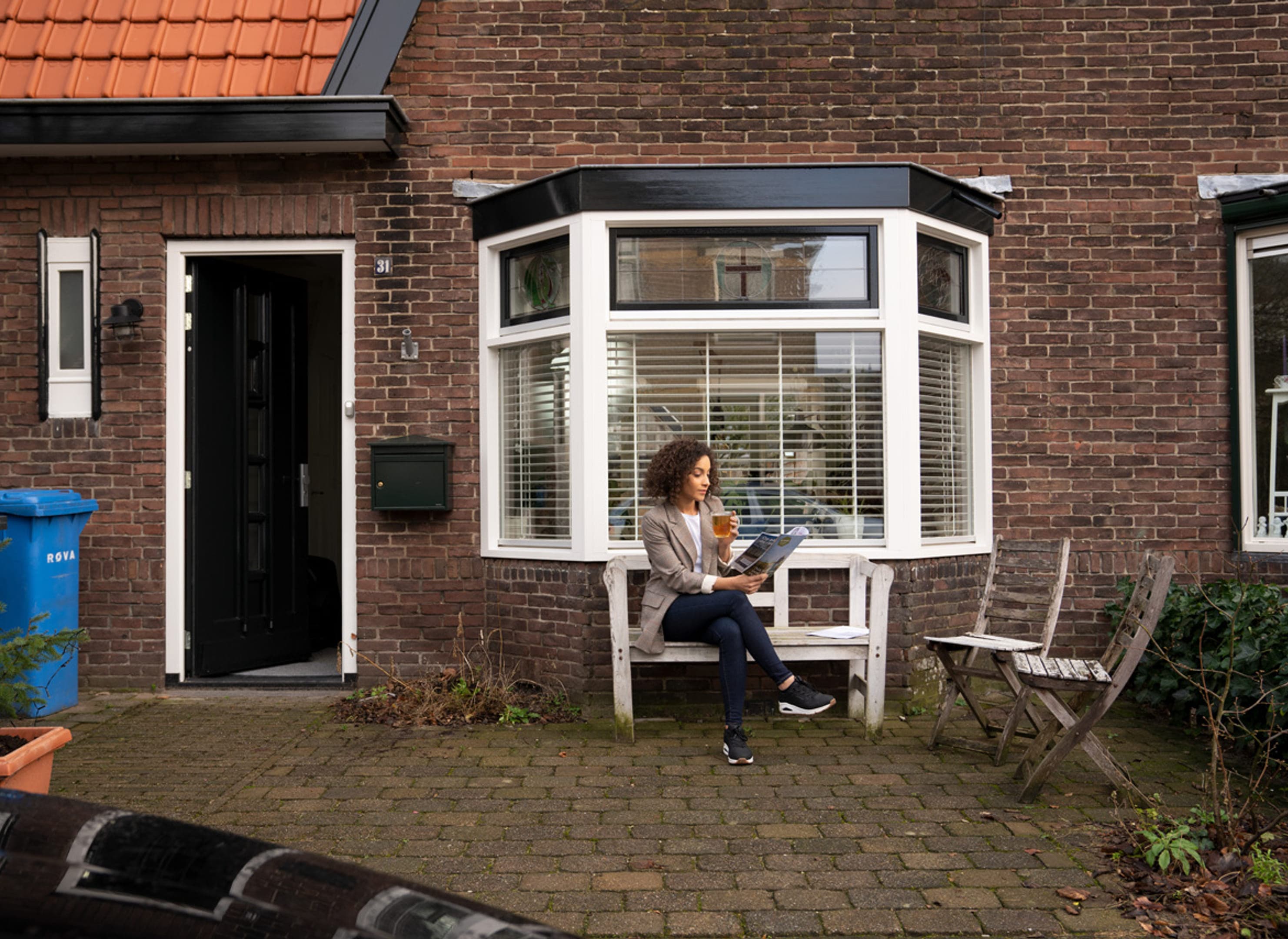 Vrouw drinkt thee op een bankje voor haar wat oudere woonhuis tegen de erker aan.