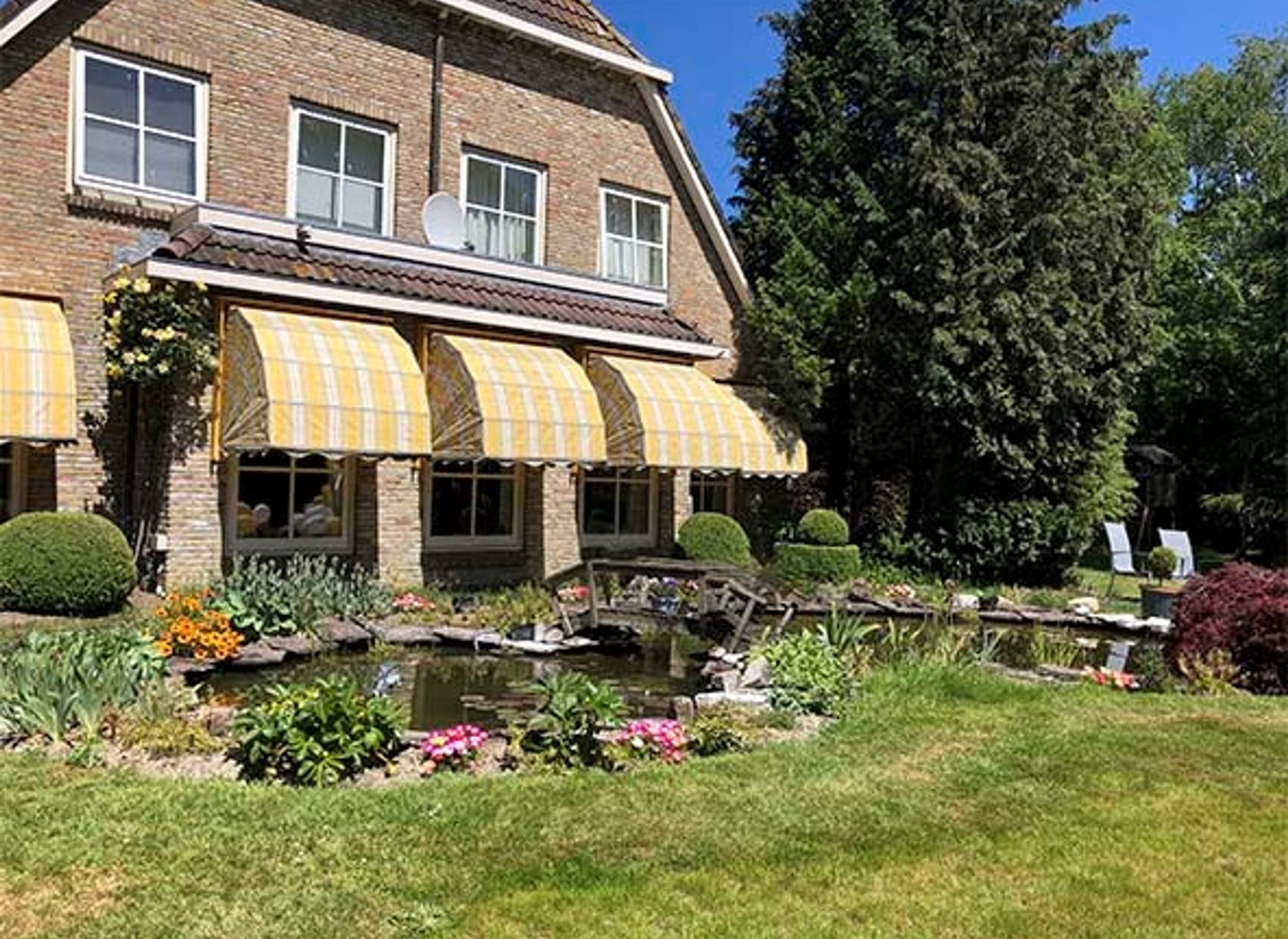 Vrijstaande woning met zonneschermen uitgeklapt, een tuin met vijver, gras en kleurrijke planten op de voorgrond