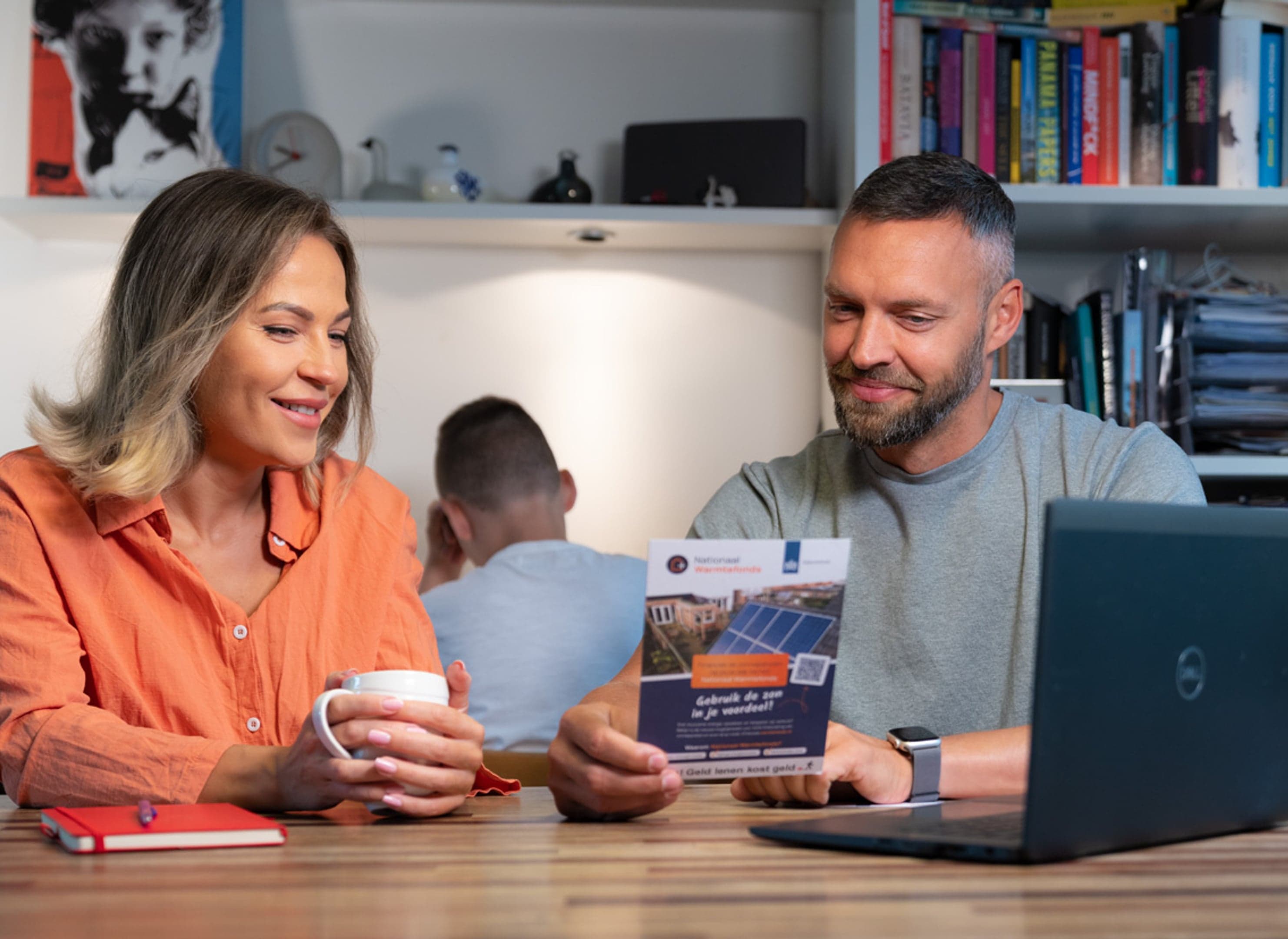 Man en vrouw bekijken samen de folder van Nationaal Warmtefonds, naast hen staat een laptop. In de achtergrond zit hun zoon met zijn rug naar hen toe aan een bureautje.