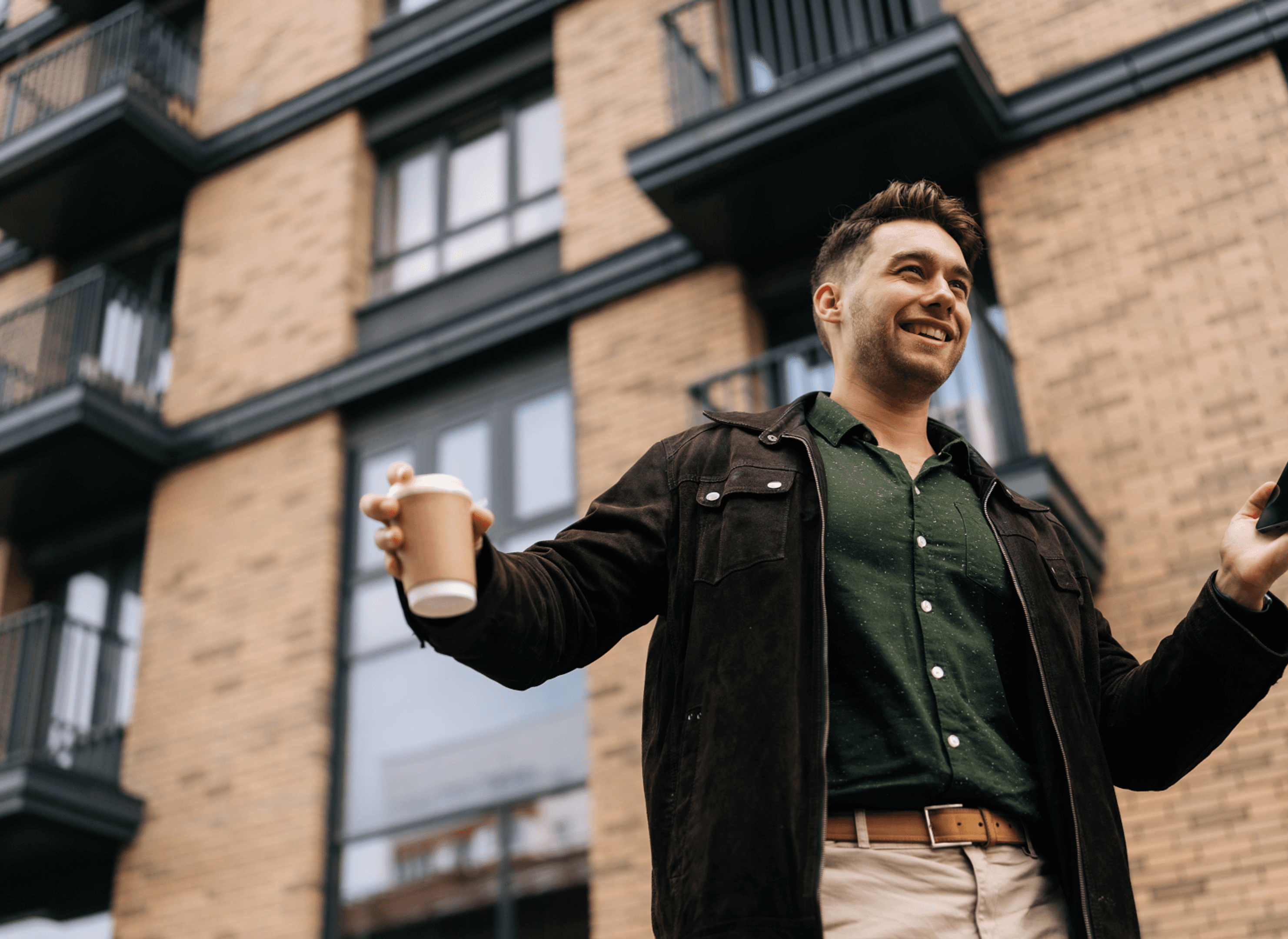 Lachende man voor een appartementsgebouw, met in zijn rechterhand een kop koffie en in zijn linkerhand zijn telefoon.