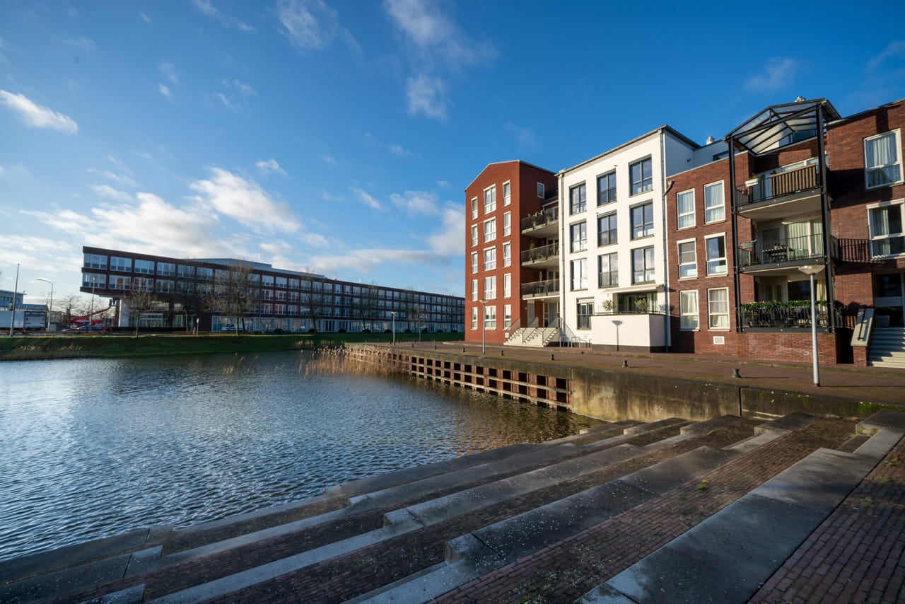 Rijtje appartementsgebouwen aan het water in rode baksteen en witte pleister, op de achtergrond een kantoorgebouw.