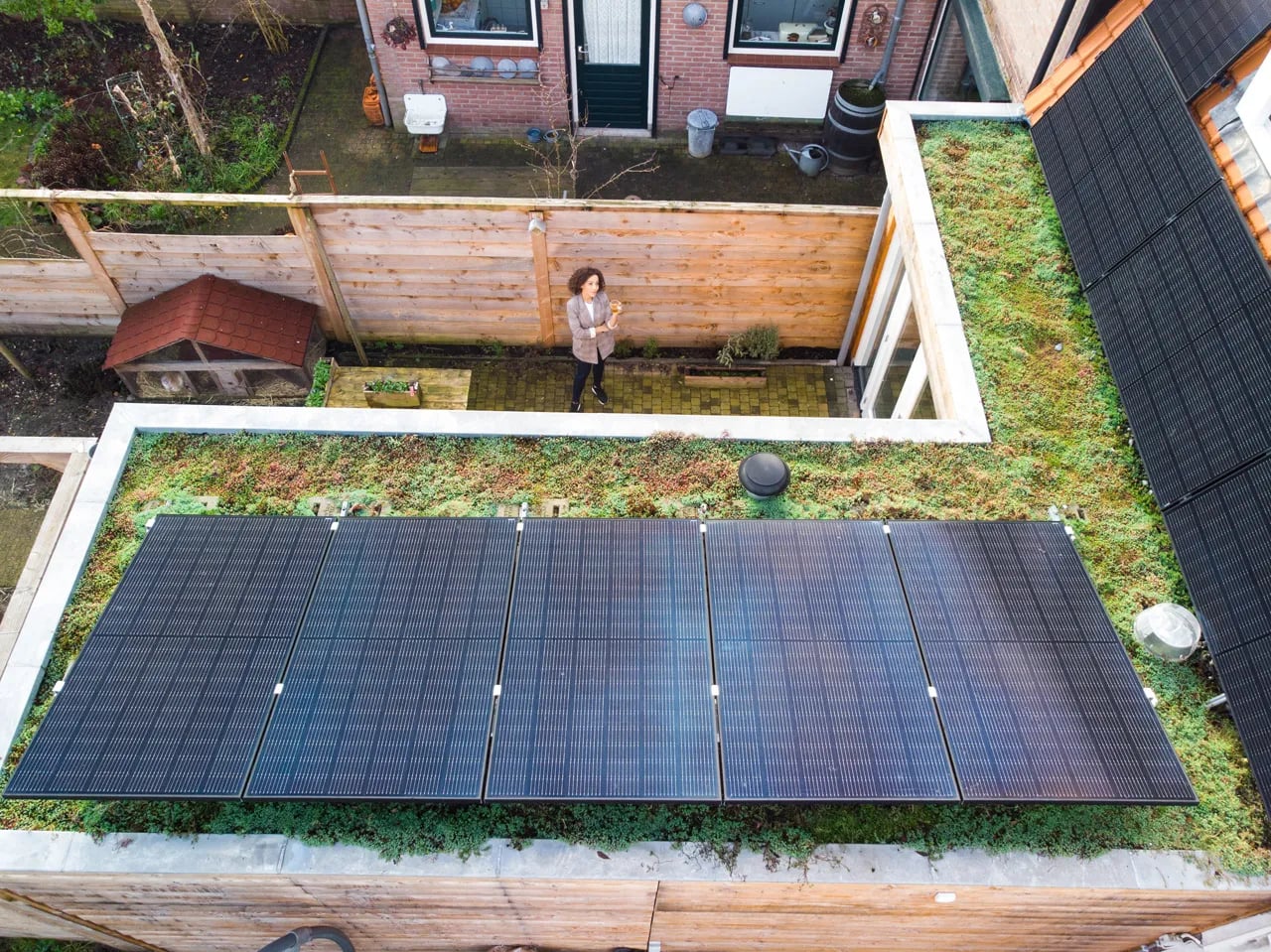Hoge drone foto, vrouw onderin met armen gespreid, lage zonnepanelen in beeld omgeven door groene daktuin.