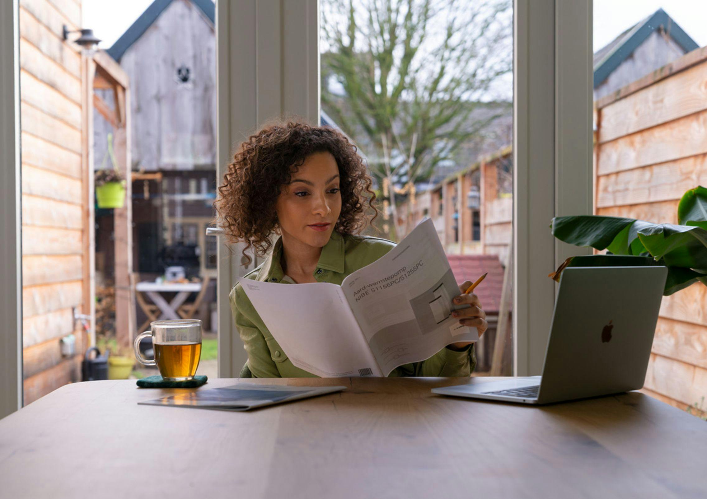Vrouw aan keukentafel, kijkend in folder, folder in beeld, titel Warmtepomp leesbaar. Tuindeuren op achtergrond, Apple laptop op tafel.