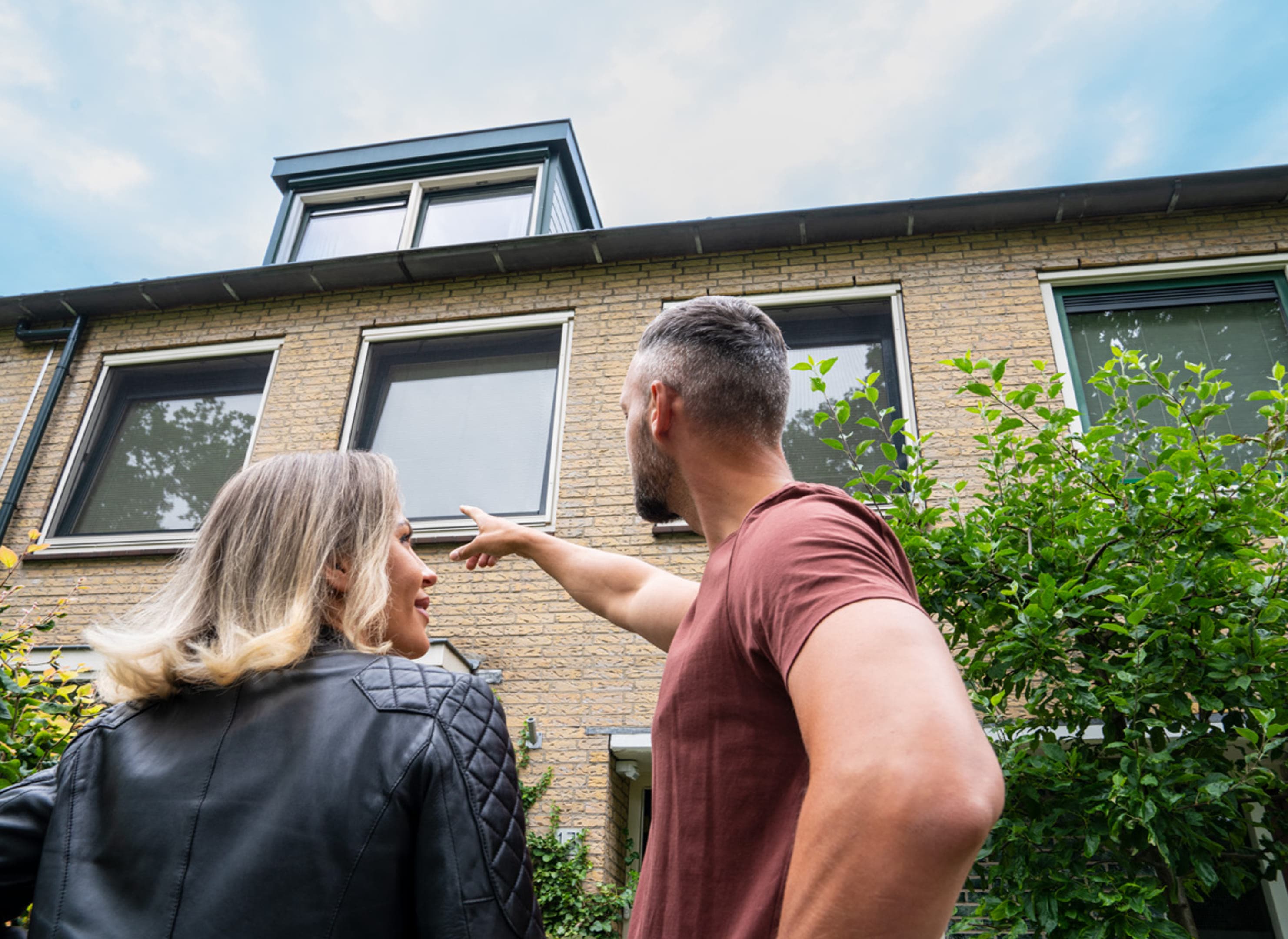 Man en vrouw voor een rijtjeshuis in geel baksteen met een kleine dakkapel, de man wijst naar de ramen op de bovenverdieping.