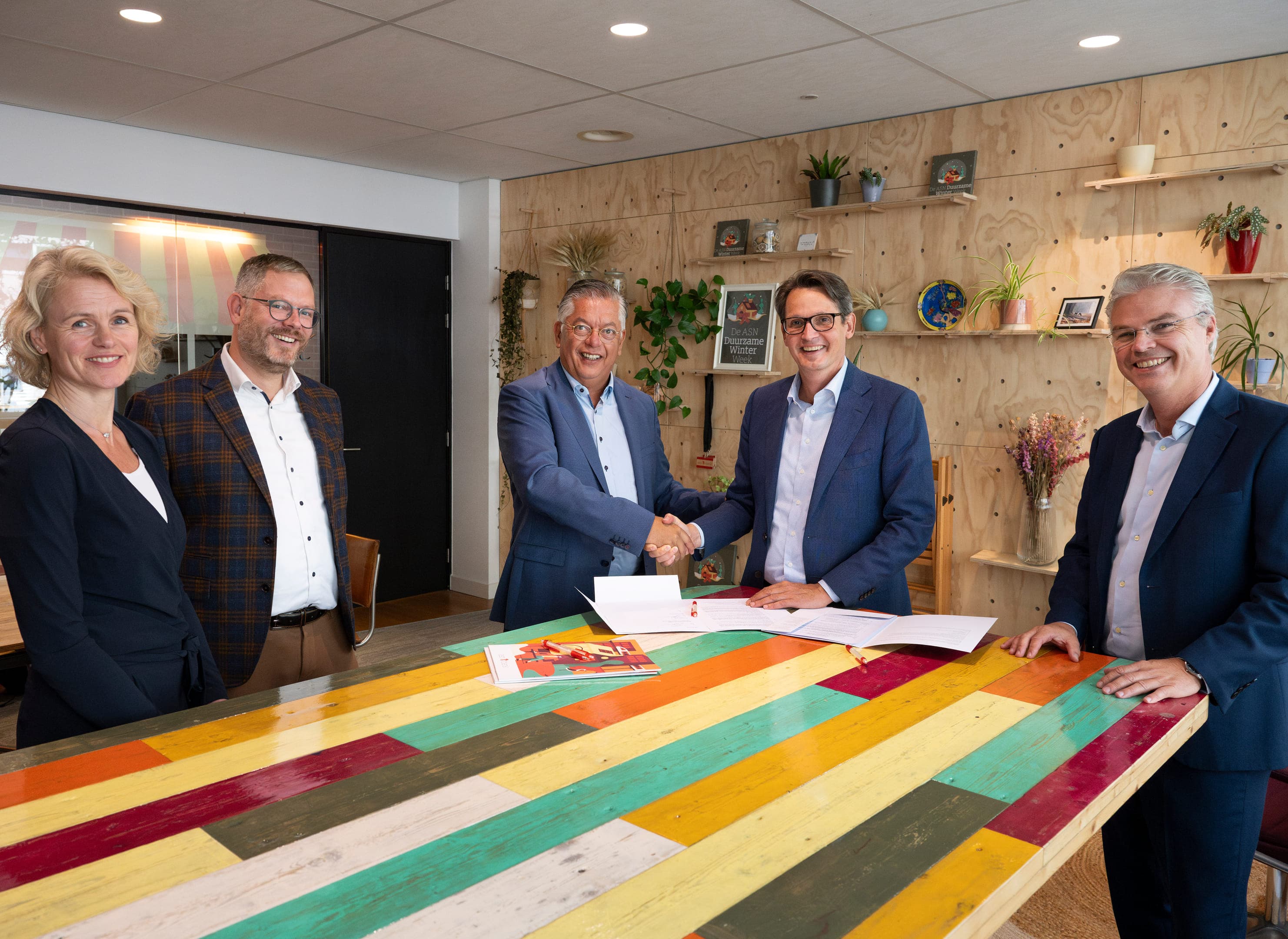 Een gezelschap van vijf mensen staat om een tafel met een felgekleurd blad, op tafel papieren die net zijn ondertekend. Het is de bekrachtiging van de samenwerking tussen ASN Bank en Nationaal Warmtefonds. Arie Koornneef van ASN Bank en Frans Overdijk van Nationaal Warmtefonds schudden elkaar de hand.