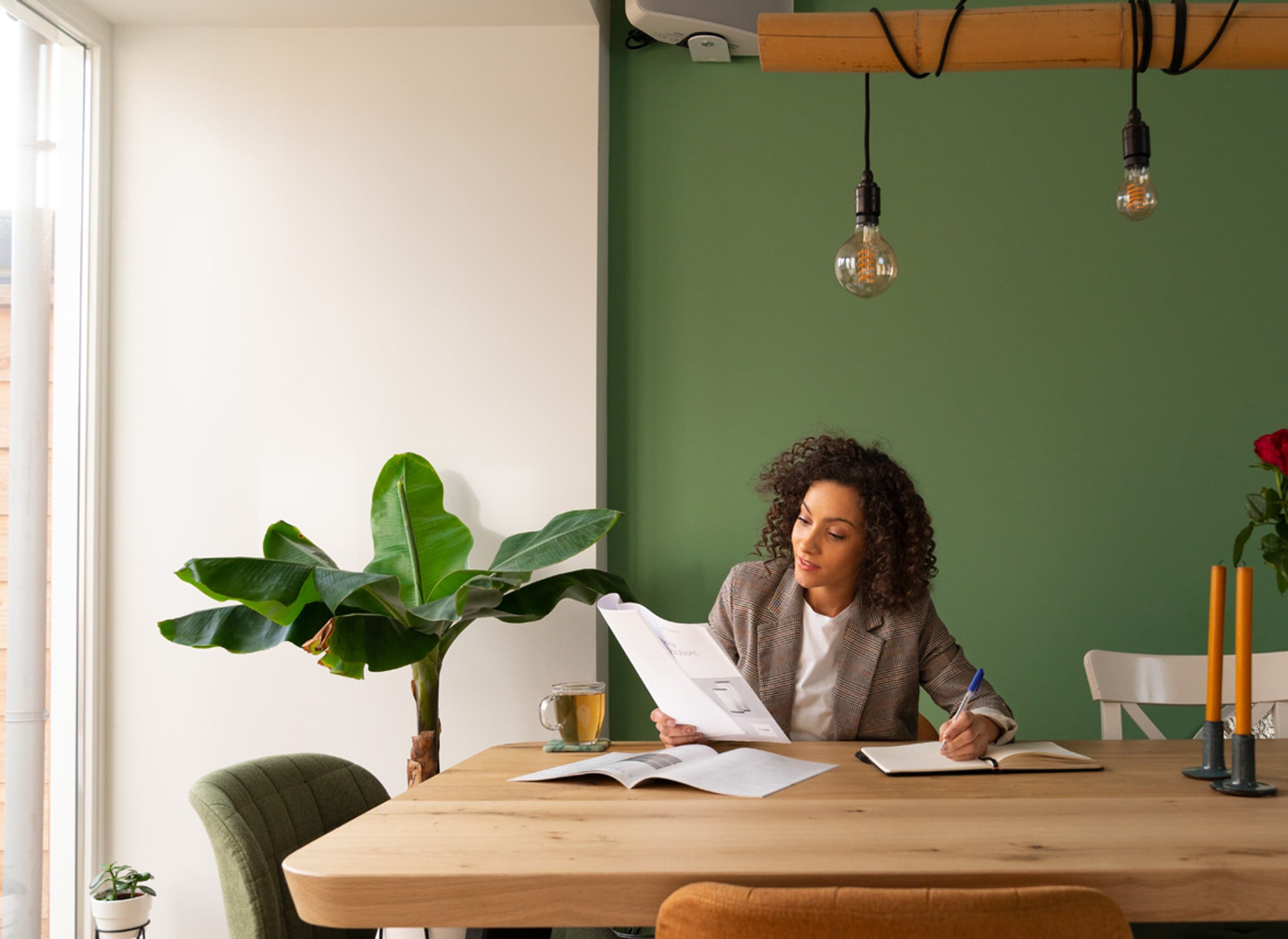 Vrouw met kop thee kijkend in folders zonder laptop, pen in hand met notitieboekje.