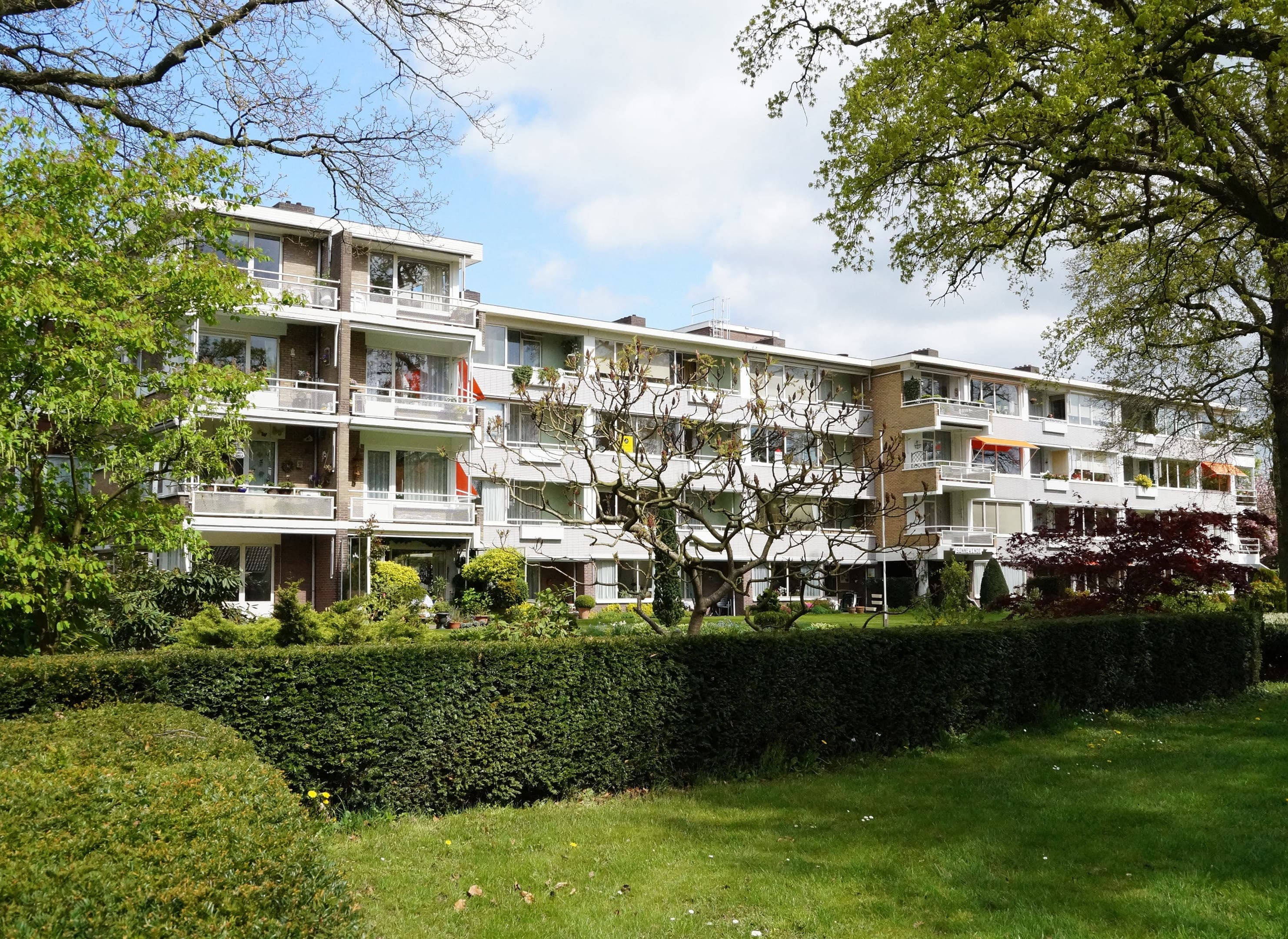 Appartementsgebouw bestaande uit drie delen, linker deel steekt naar voren uit. Op de voorgrond gras en een heg.